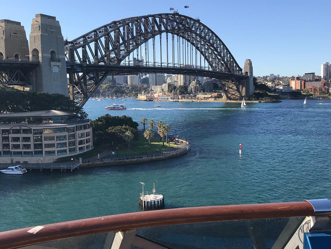 Sydney Harbour Bridge over blue water, with boats, shoreline buildings, and a railing in the foreground.