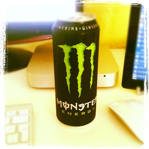 Black Monster Energy drink can on a desk between a keyboard and mouse, with a computer and monitor in the background.