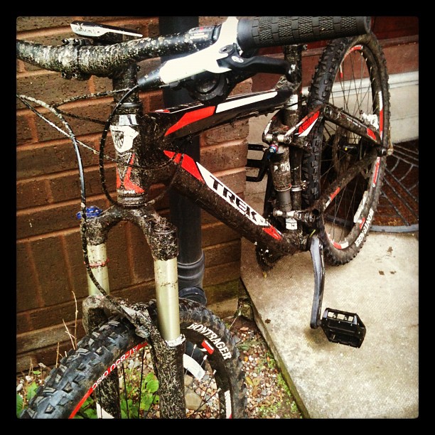 Mud-covered black and red mountain bike leaning against a brick wall beside a concrete step.