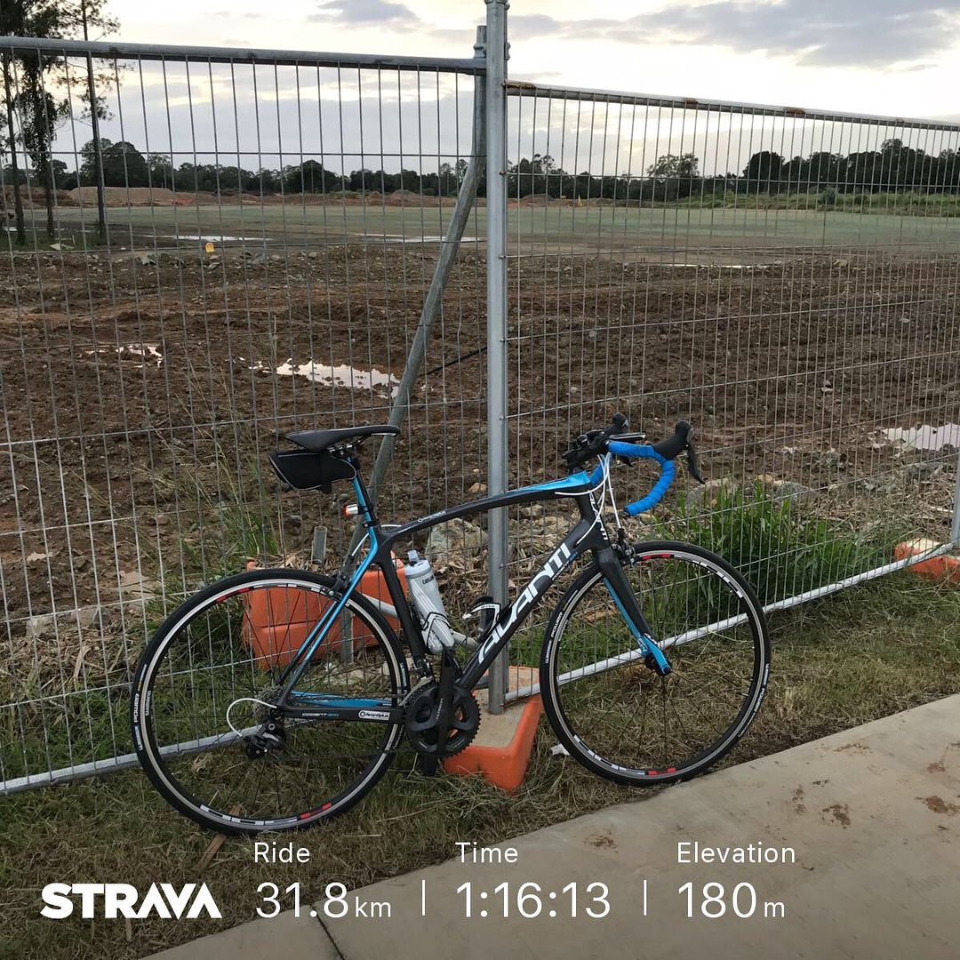 Black and blue road bike leaning against a metal fence beside a muddy field, with Strava stats reading 31.8 km, 1:16:13, and 180 m.