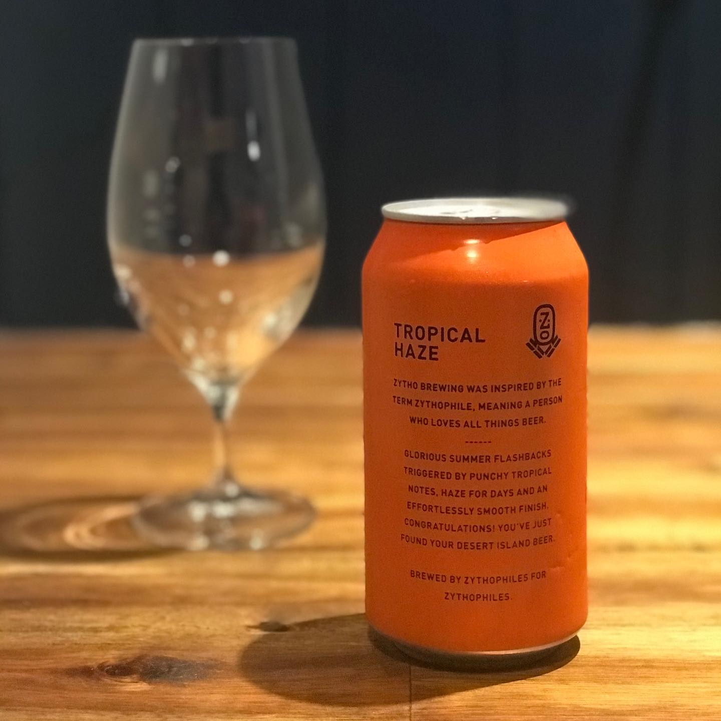 Orange Tropical Haze beer can on a wooden table beside a partially filled stemmed glass against a dark background.