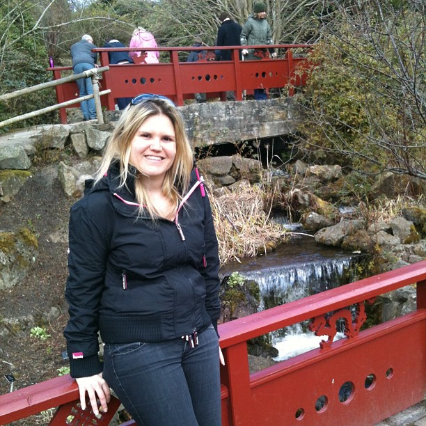 Woman in a black jacket standing by a red railing, with a rocky stream and people on a red bridge behind her.