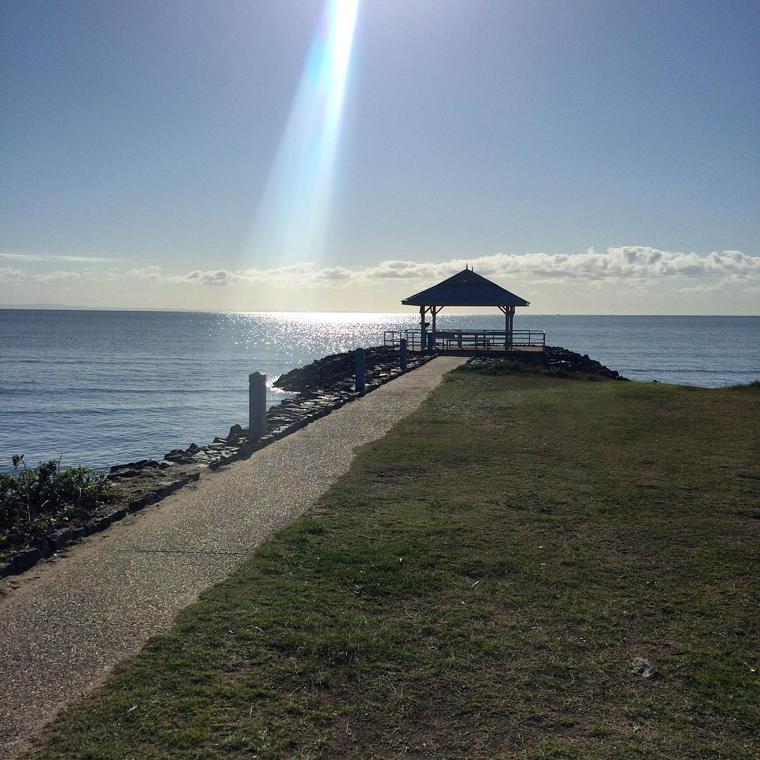 Path leading to a small gazebo on a rocky point beside calm water, with bright sun glare and a rainbow-like beam in the sky.