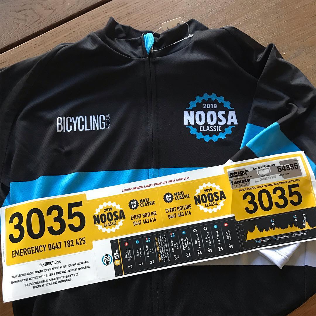 Black cycling jersey with blue accents and 2019 Noosa Classic logos, laid on a table with race bib number 3035 and RFID timing tag.