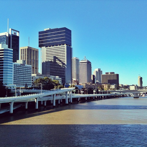 City skyline with modern high-rise buildings beside a river, crossed by an elevated roadway under a clear blue sky.