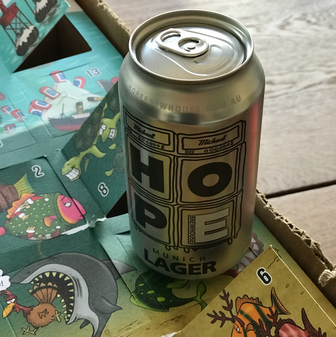 Silver can labeled "HOPE Munich Lager" standing on a colorful illustrated cardboard surface.