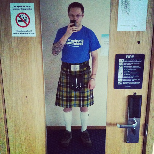 Person taking a mirror selfie in a blue T-shirt, tartan kilt, white knee socks, and shoes inside a doorway.
