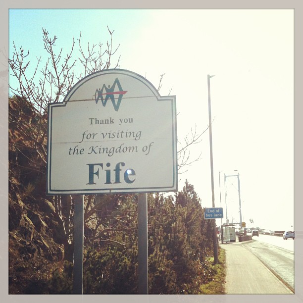 Roadside sign reading "Thank you for visiting the Kingdom of Fife" beside a sunlit road with bare trees and streetlights.