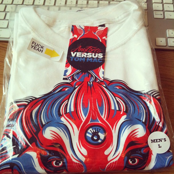Folded white T-shirt in plastic packaging with a red and blue face graphic, a central tag, and a “MEN’S L” sticker.