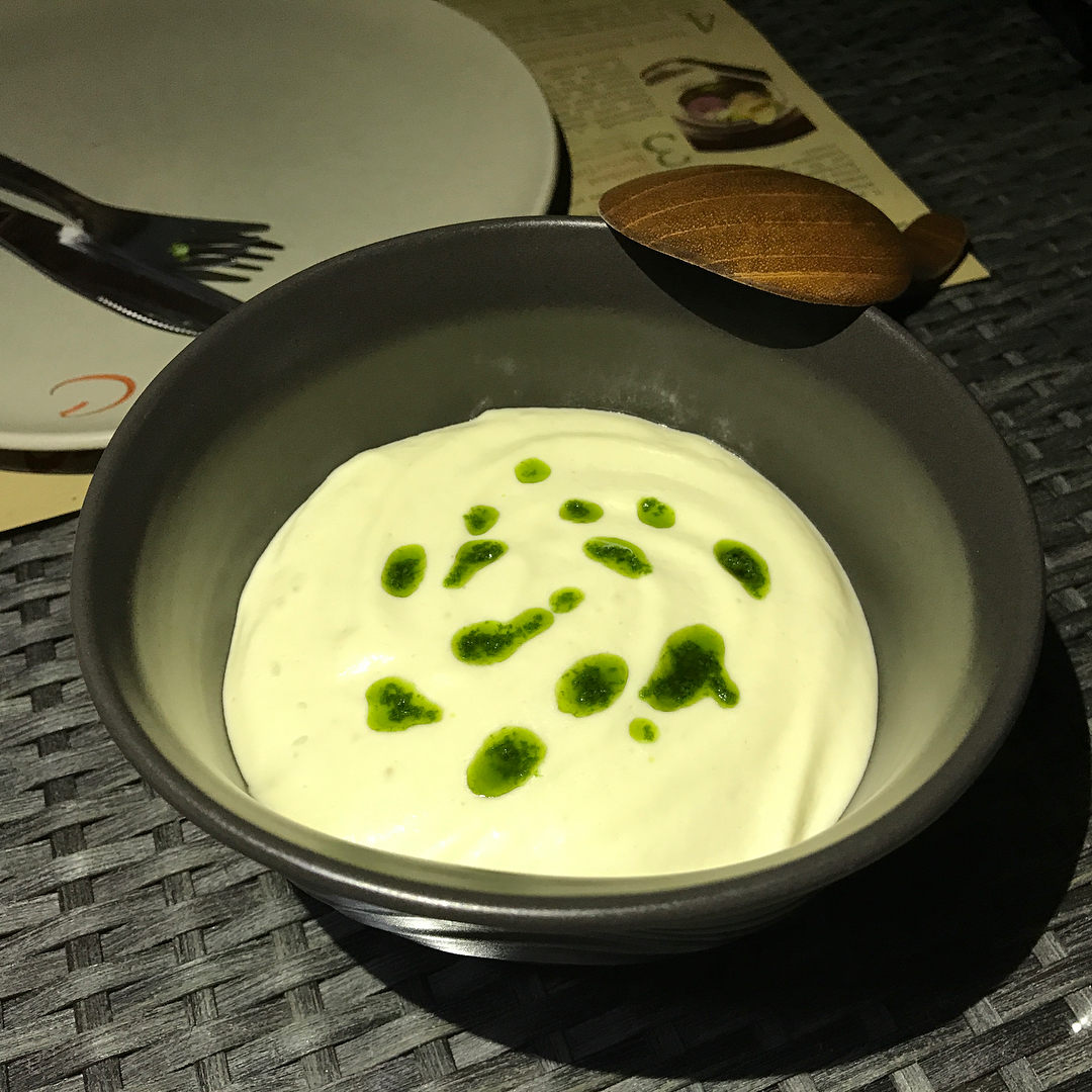 Bowl of creamy white soup or puree topped with green herb oil, with a wooden spoon and plate with fork in the background.