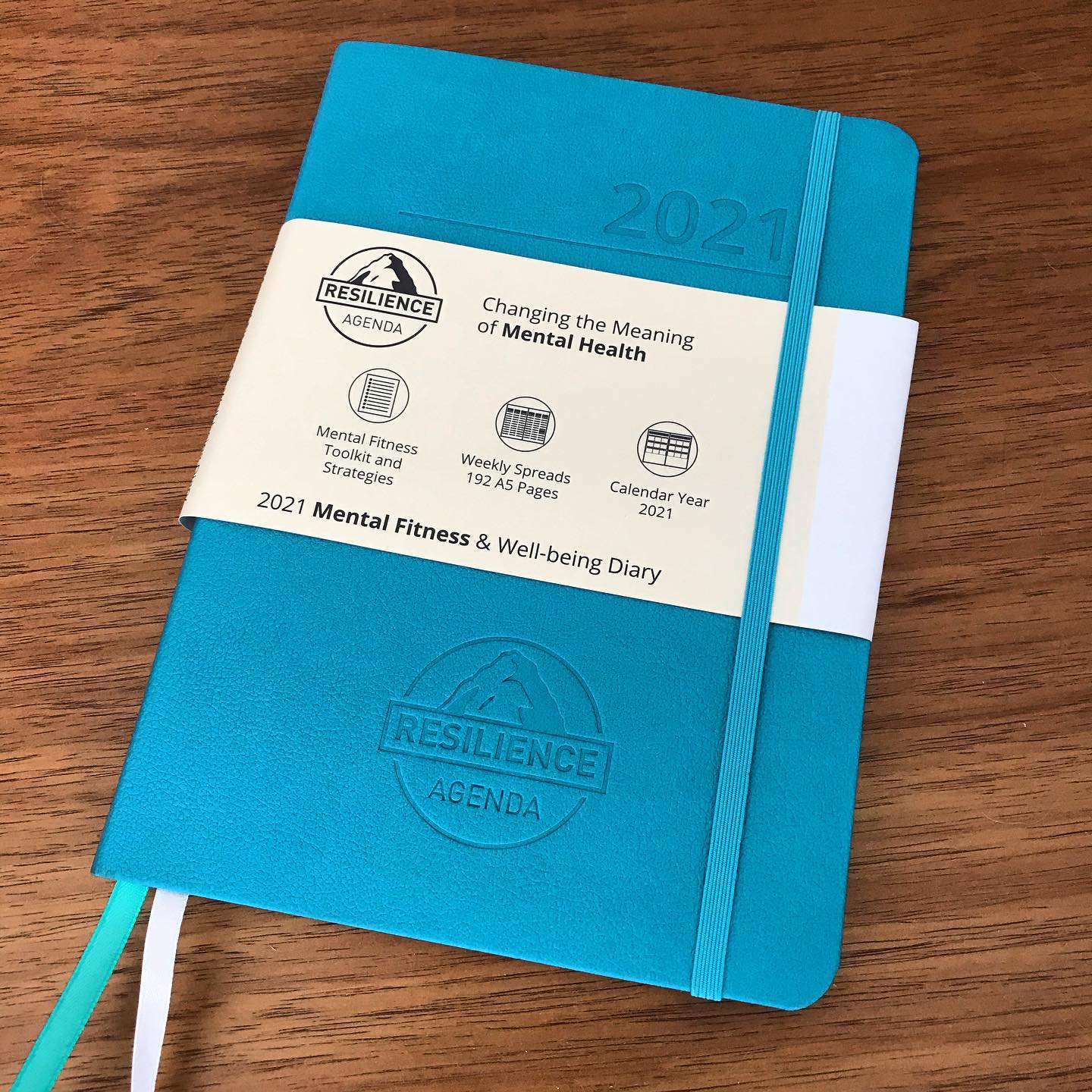 Blue 2021 Resilience Agenda mental fitness and well-being diary with a white paper band, lying on a wooden table.