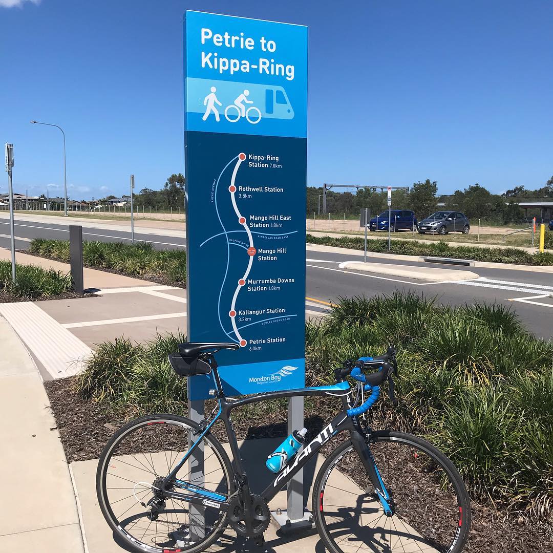 Road bike parked beside a blue Petrie to Kippa-Ring path sign showing station stops and distances on a sunny day.