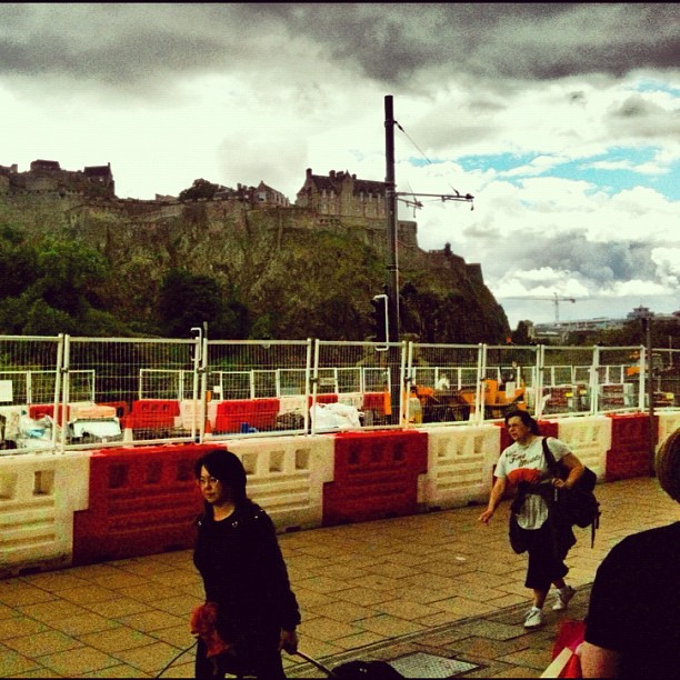 People walk with luggage past a fenced construction area, with a rocky hilltop castle under dark clouds in the background.