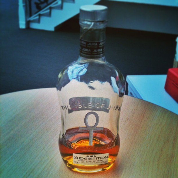 Partially empty Jura Superstition whisky bottle with an ankh symbol on the front, standing on a round wooden table.