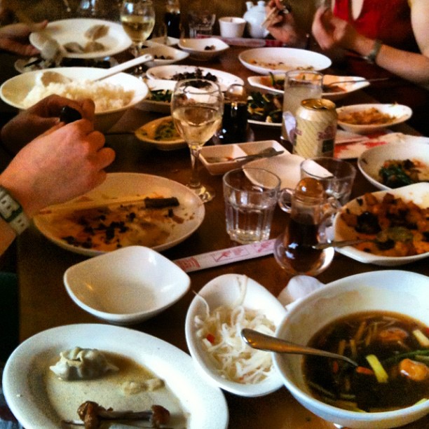 People eating at a crowded restaurant table covered with plates, bowls, drinks, and leftover food.