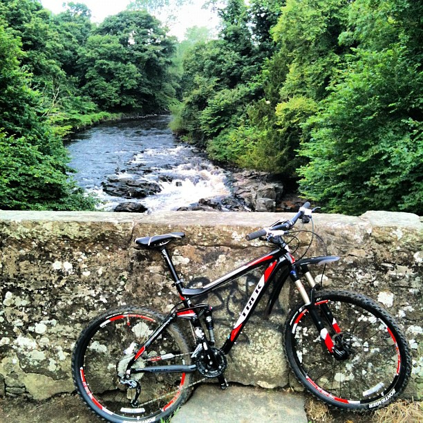 Black and red mountain bike leaning against a stone bridge wall above a rocky river surrounded by dense green trees.