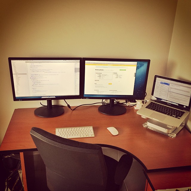Desk setup with two monitors, a laptop on a stand, keyboard, mouse, and office chair against a plain wall.