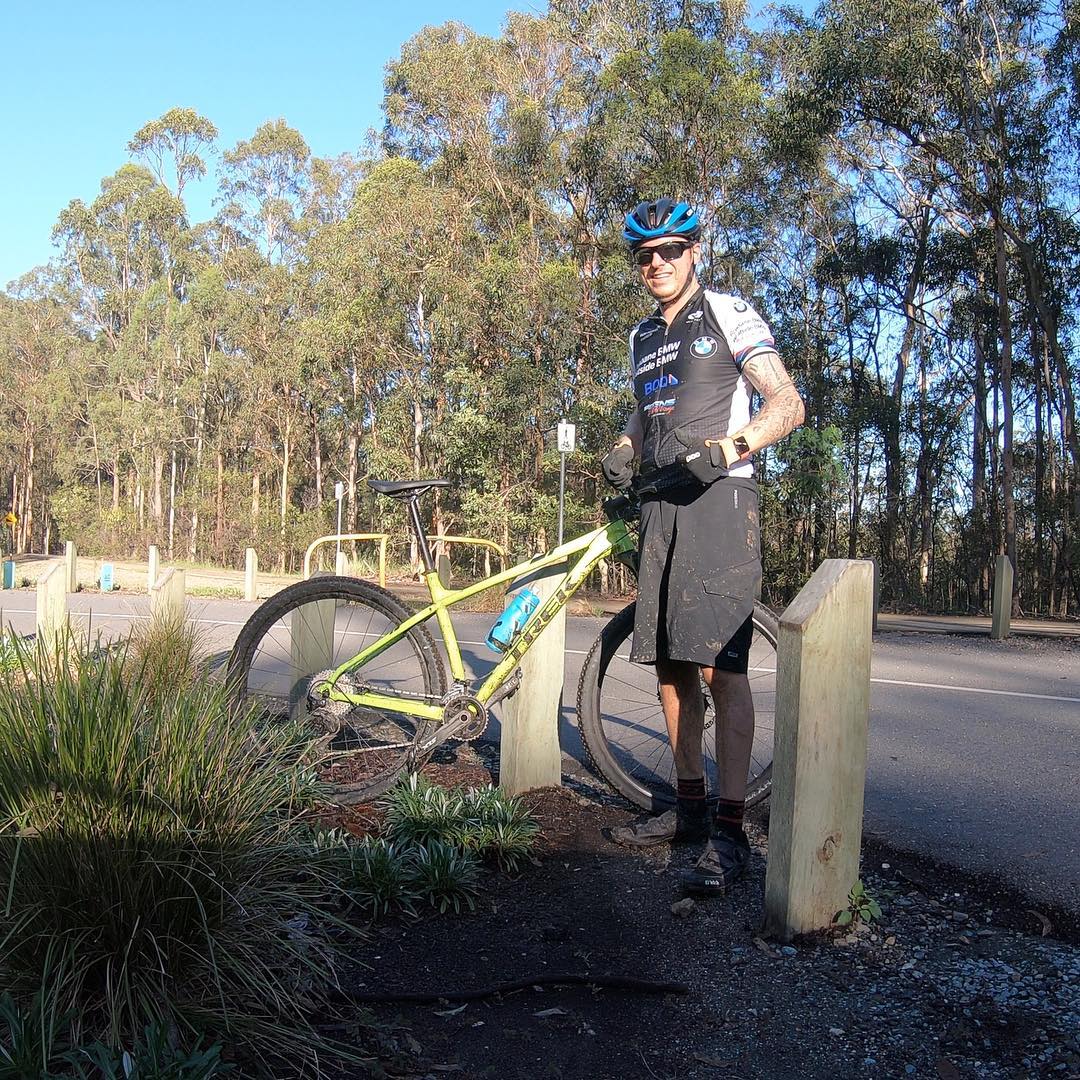 Cyclist in a blue helmet and black jersey stands beside a yellow mountain bike near a roadside path with tall trees in the background.