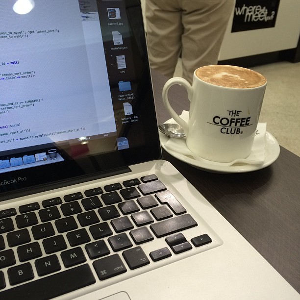 MacBook Pro beside a cappuccino in a white Coffee Club cup on a saucer, with a person standing in the background.