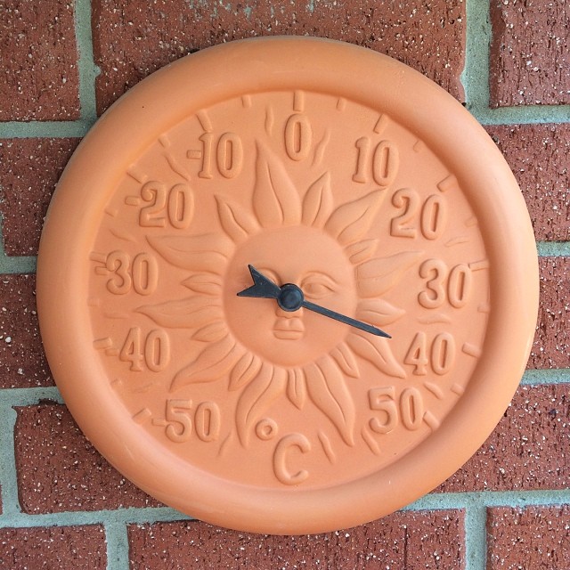 Terracotta wall thermometer with a sun face design mounted on a red brick wall.