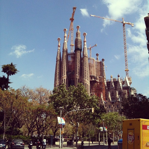 Cathedral with tall spires under construction, surrounded by cranes, trees, and a street with traffic lights and parked scooters.