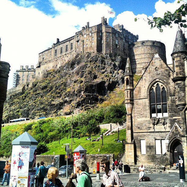 Stone castle on a rocky hill above a street, with a church building in the foreground and people walking below.