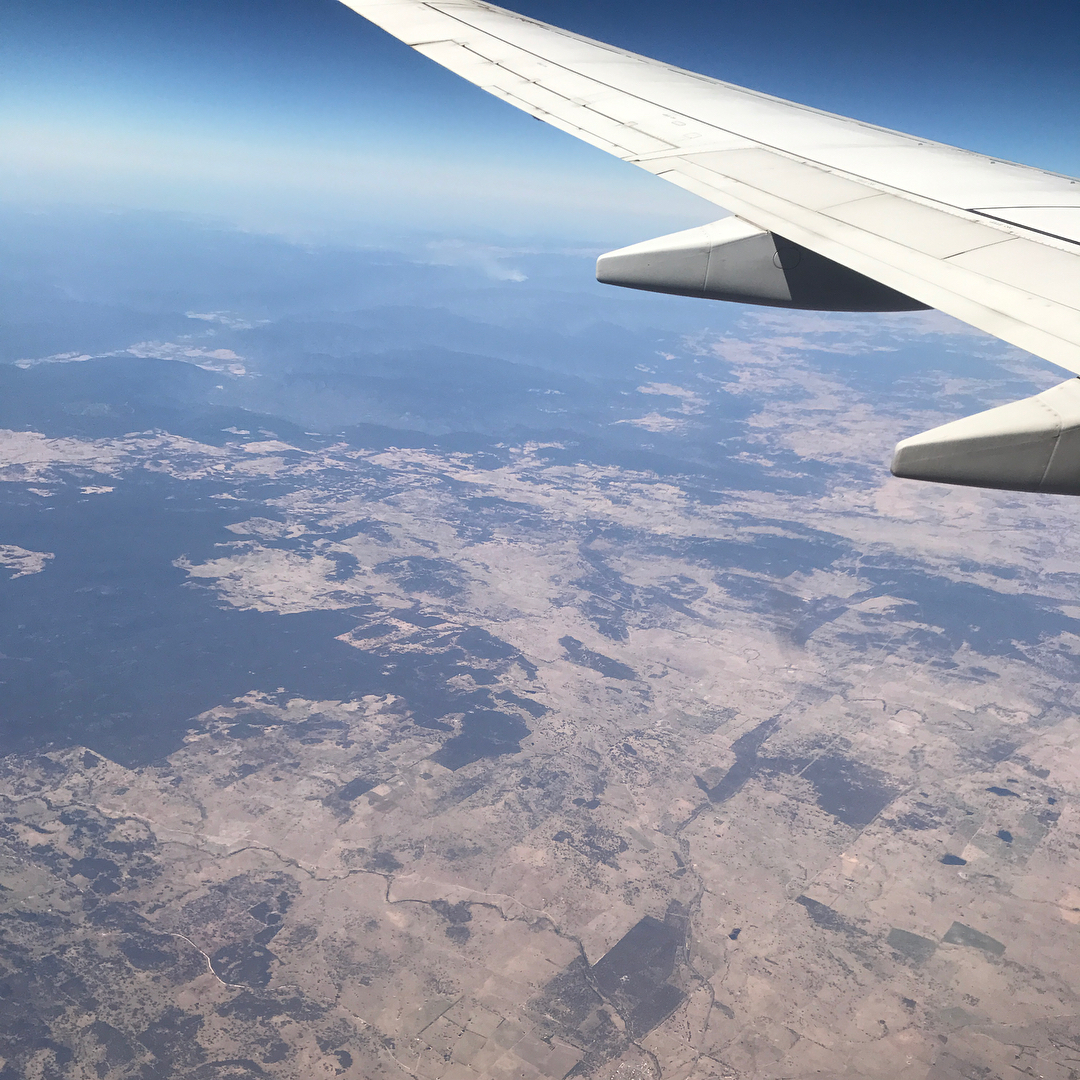 View from an airplane window showing a wing above a dry patchwork landscape under a clear blue sky.