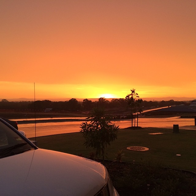 Orange sunset over a river and grassy bank, with silhouetted trees and the front corner of a white car in the foreground.