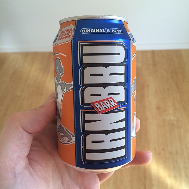 Hand holding a can of Barr IRN-BRU with orange and blue label against a wooden floor background.