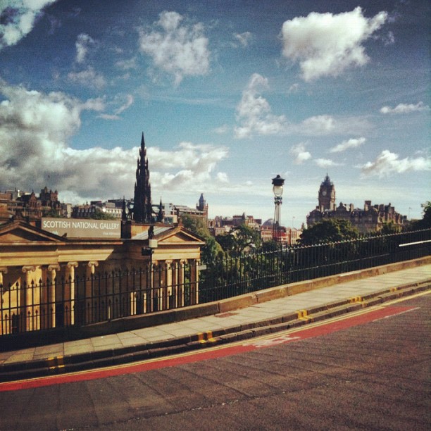 View of Edinburgh skyline with the Scottish National Gallery, church spire, streetlamp, and curved road under a partly cloudy sky.