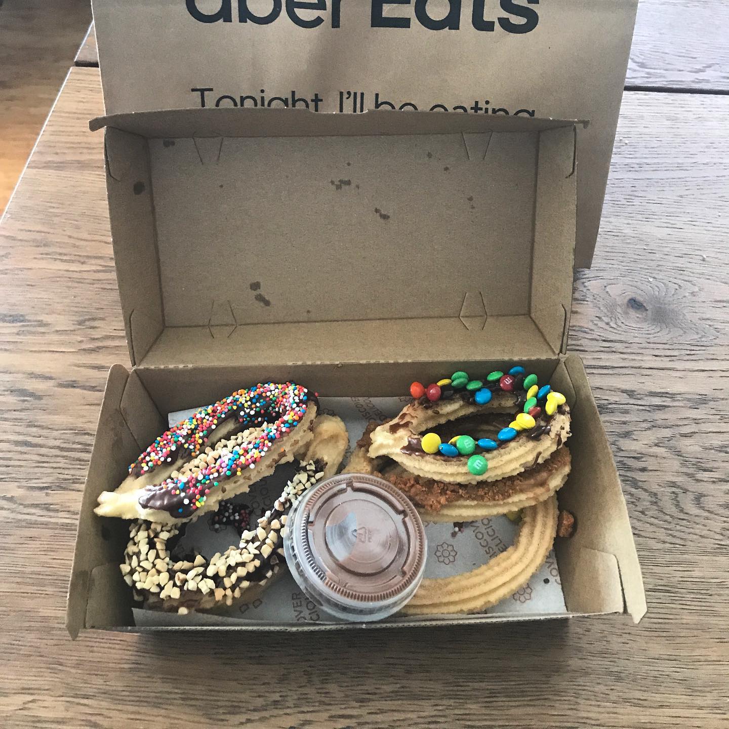 Open cardboard box with assorted chocolate-dipped churros topped with sprinkles, chopped nuts, and candy-coated chocolates beside dipping sauce.