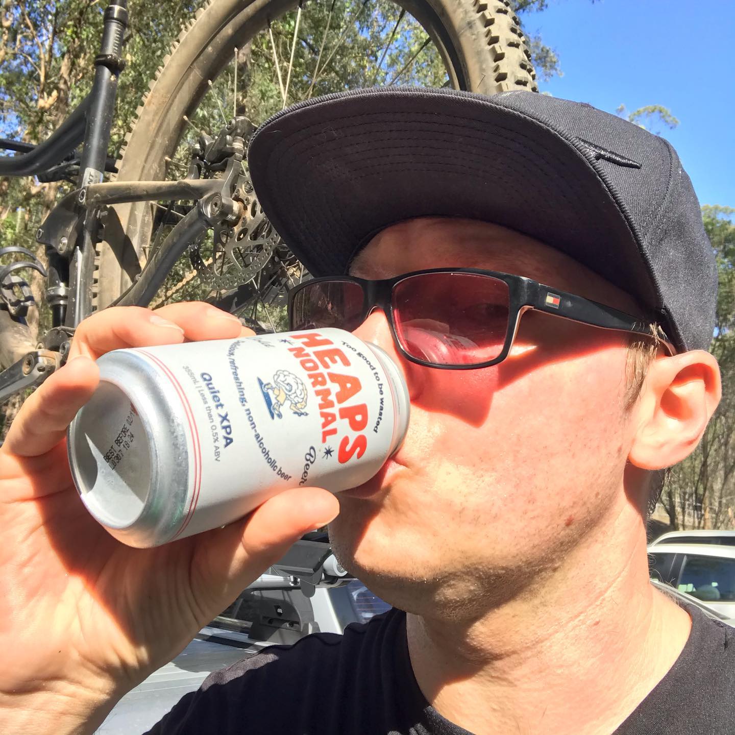 Person in a black cap and sunglasses drinks from a can beside a mountain bike mounted on a car rack in a sunny wooded area.