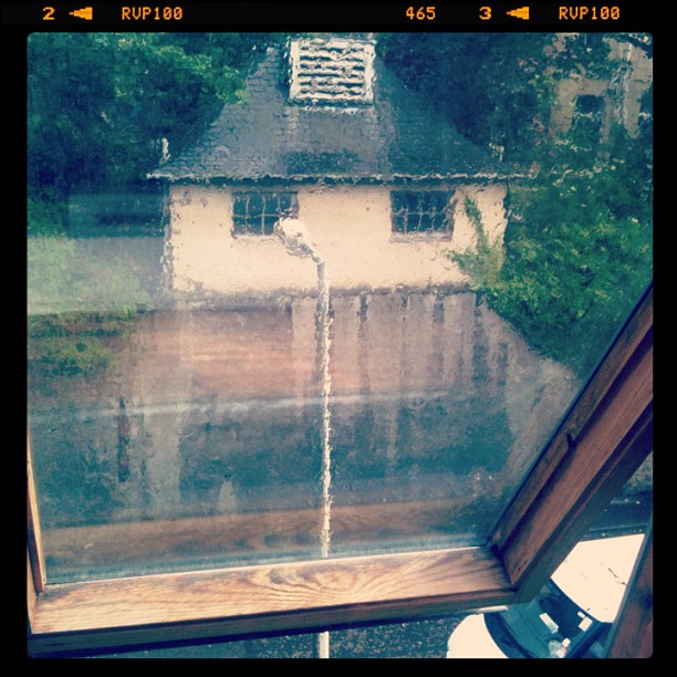 Rain-streaked skylight framing a blurred view of a small white building and wet driveway surrounded by trees.