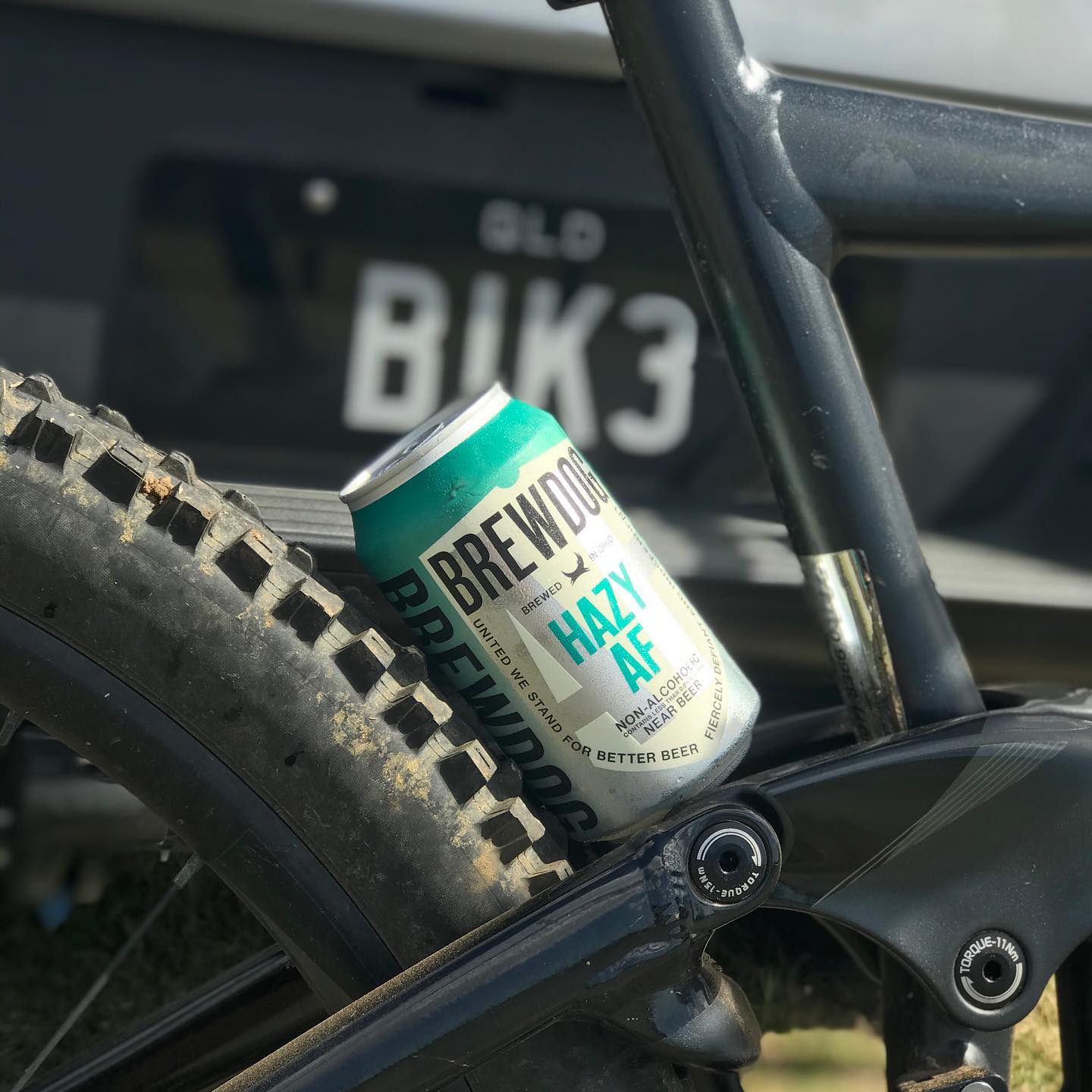 Can of BrewDog Hazy AF wedged in a mountain bike frame beside a muddy rear tire, with a blurred license plate in the background.