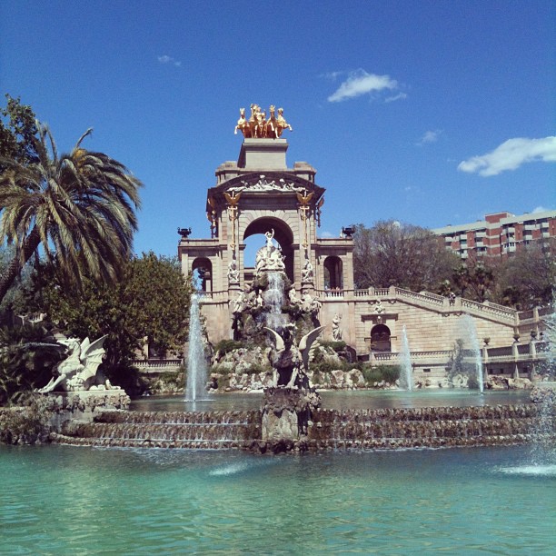 Ornate fountain with statues and water jets before a grand pavilion, with palm trees and a bright blue sky.