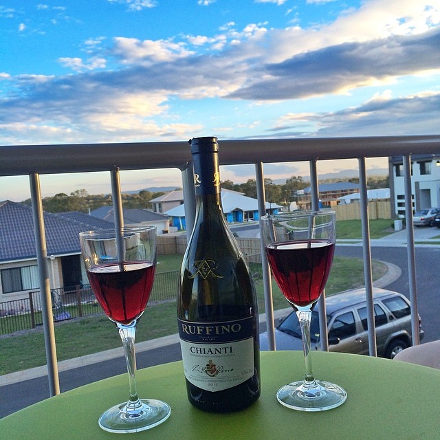 Bottle of Ruffino Chianti between two glasses of red wine on a balcony table overlooking a suburban street at sunset.