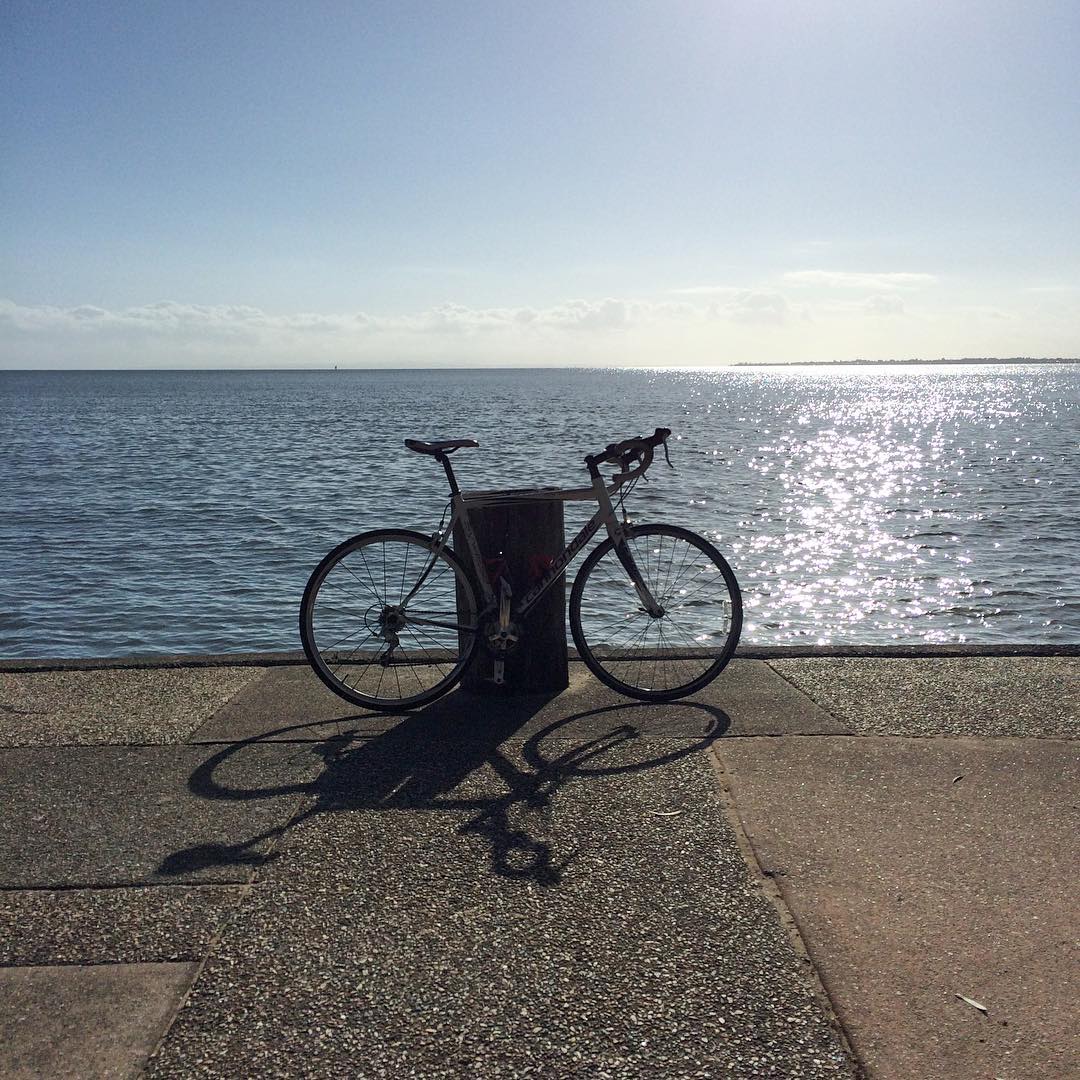 Bicycle leaning against a post on a waterfront path, silhouetted against sunlit water under a clear sky.