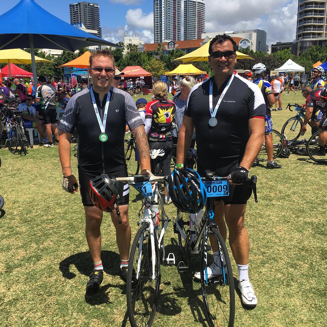 Two cyclists wearing medals stand with their road bikes on a grassy field among event tents and other riders, with tall buildings behind.