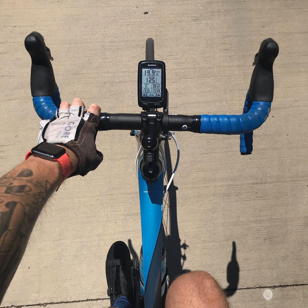View from a cyclist riding a blue road bike, with one gloved hand on the handlebar and a bike computer centered above the stem.