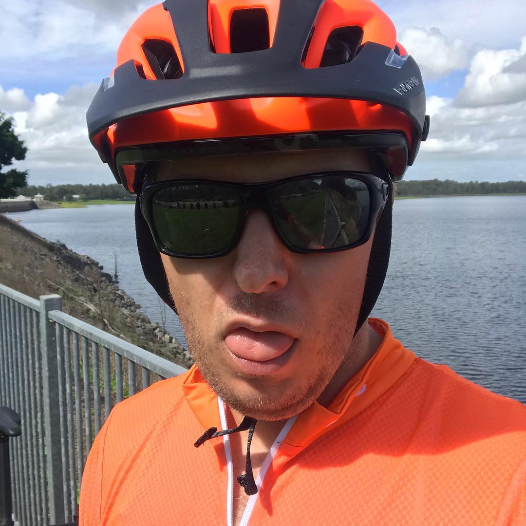 Person in an orange bike helmet and jersey wearing sunglasses, sticking out their tongue beside a body of water under a cloudy sky.