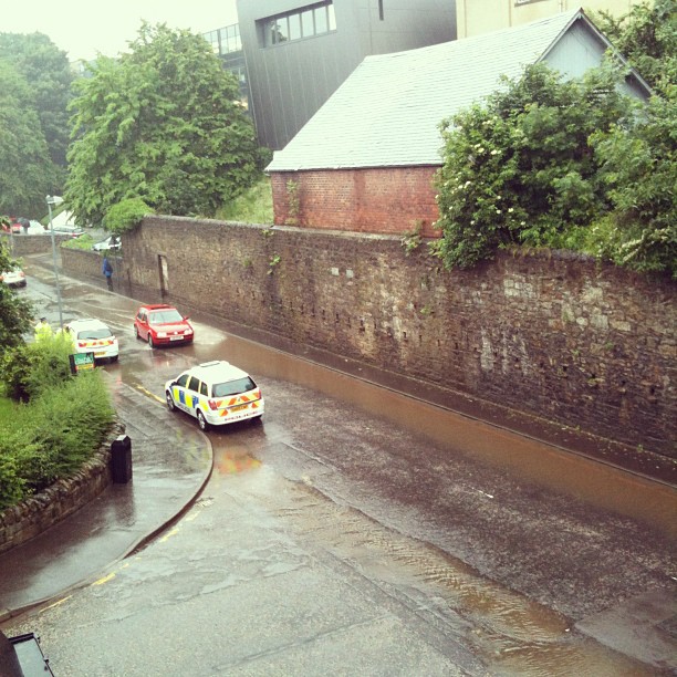 Rain-soaked street beside a high stone wall, with three cars driving through shallow floodwater.