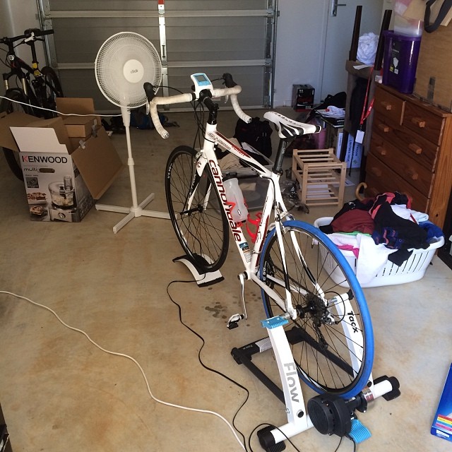 White road bike on an indoor trainer in a garage, with a standing fan, boxes, dresser, and laundry basket nearby.