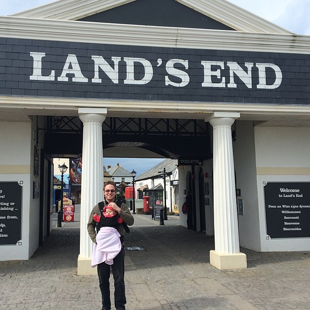 Person carrying a baby stands between white columns beneath a large Land’s End sign at an entranceway.