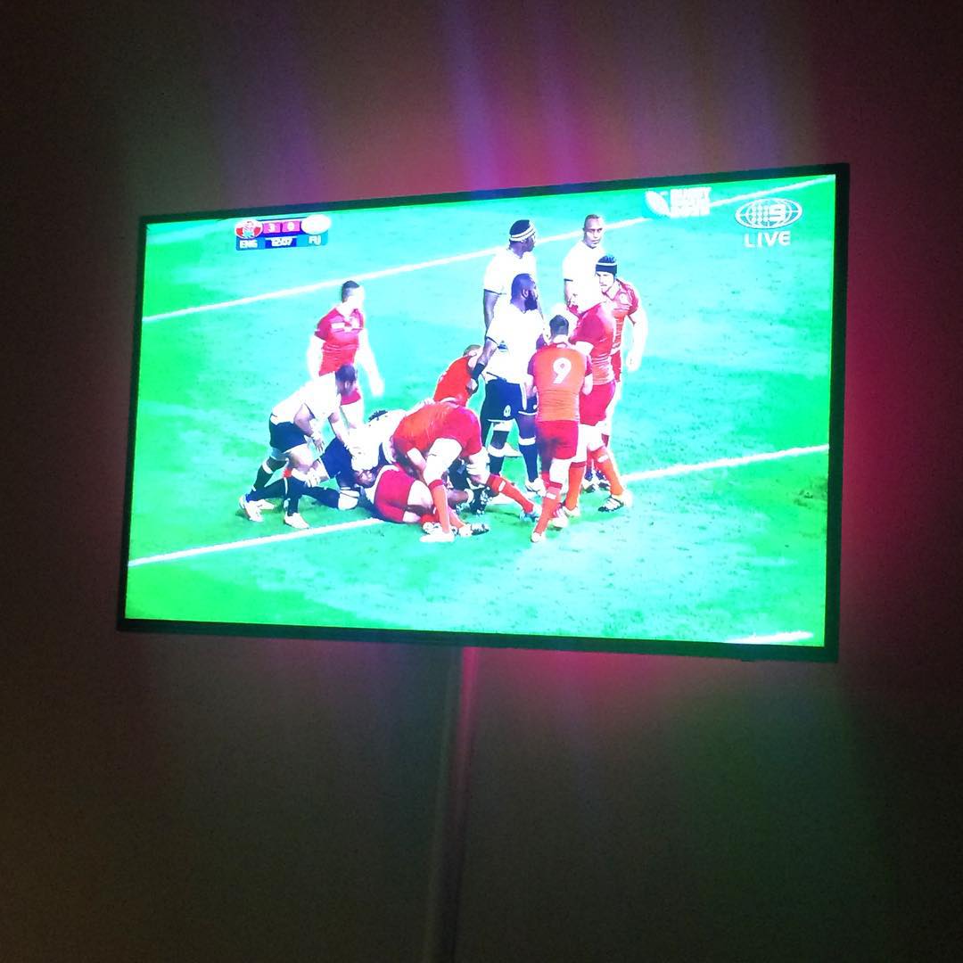 Television showing a live rugby match, with players in red and white gathered in a tackle on a bright green field.