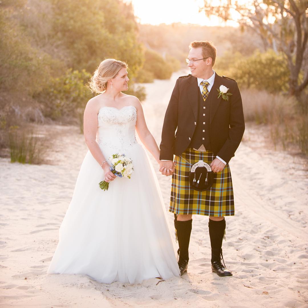 Bride in a white gown and groom in a tartan kilt hold hands on a sandy path at sunset, with the bride holding a small bouquet.
