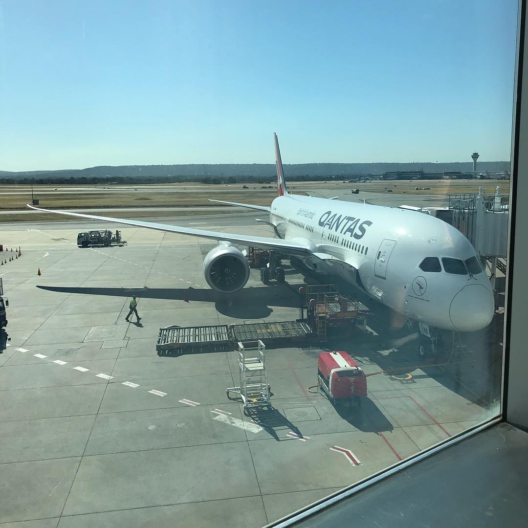 Qantas airplane parked at an airport gate, viewed through a terminal window, with ground crew and service vehicles on the tarmac.