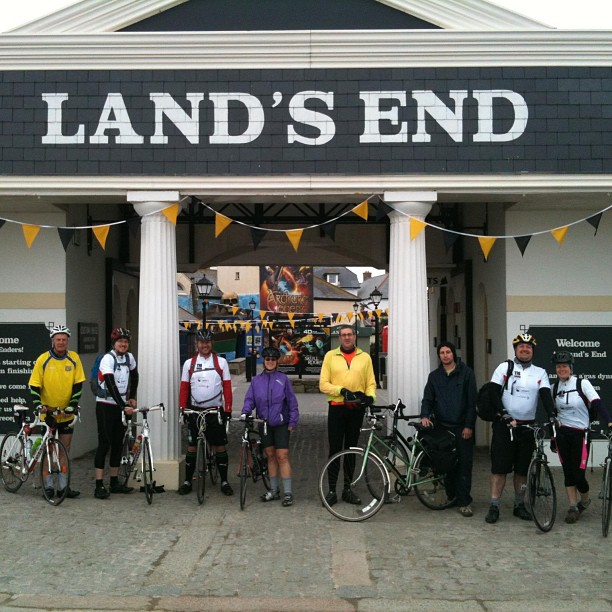 Seven cyclists with bikes stand under a large LAND’S END sign between white columns and yellow bunting.