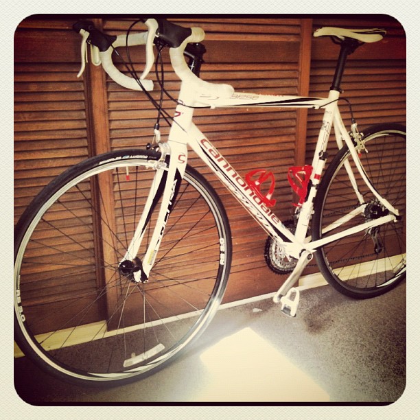 White road bicycle with drop handlebars and two red bottle cages leaning against wooden closet doors.