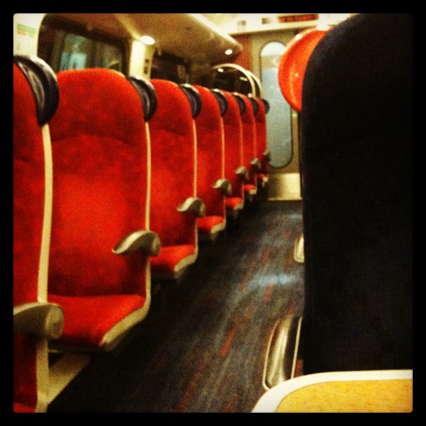 Row of red seats inside a train carriage with a dark aisle and a door at the far end.