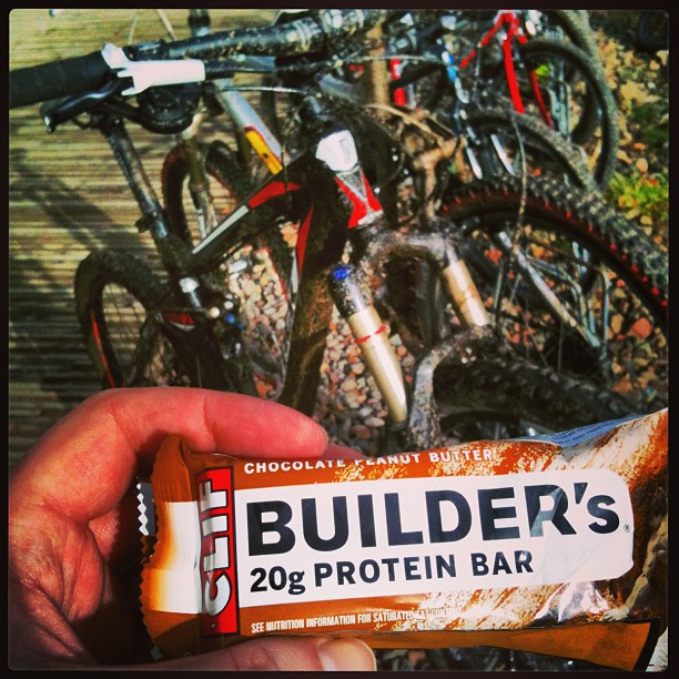 Hand holding a Builder’s chocolate peanut butter protein bar in front of several mountain bikes.